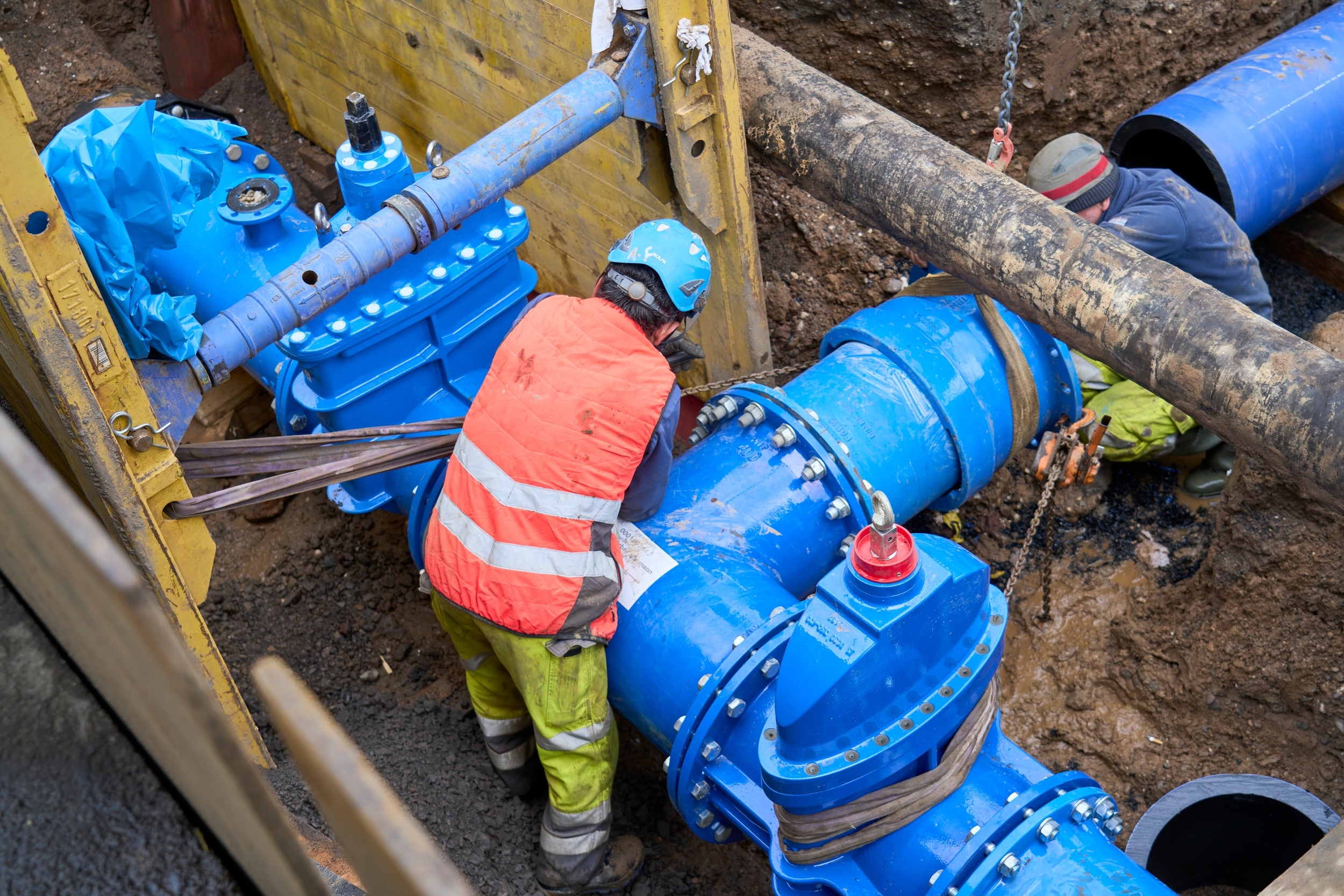 Arbeiter arbeiten an großen blauen Wasserrohren in einer Baustelle. Sie tragen Helme und Sicherheitskleidung. Die Umgebung ist eine tief gegrabene Erde, gesichert mit Holzstützen.
