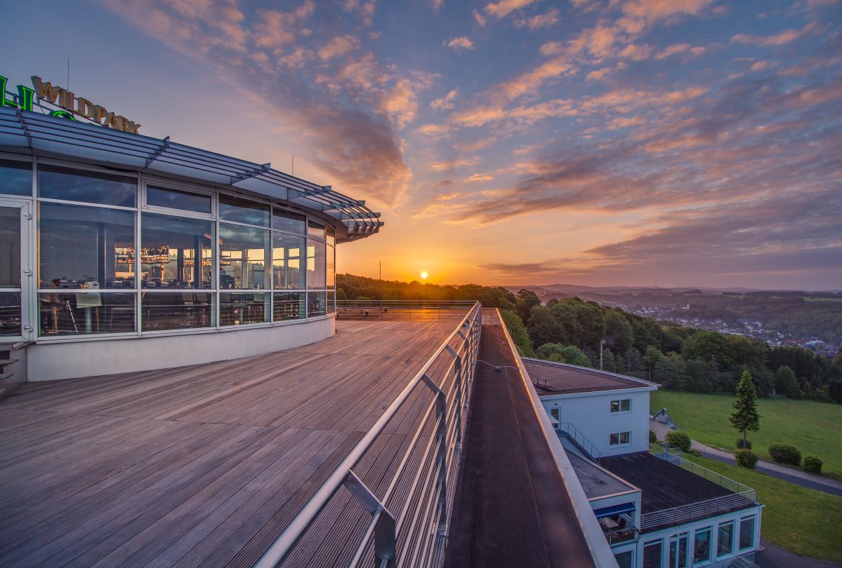 Das Wildparkhotel in Bad Marienberg Ein modernes Gebäude mit großer Terrasse überblickt bei Sonnenuntergang eine hügelige Landschaft. Im Hintergrund erstrecken sich Wälder und ein kleiner Ort unter einem farbenprächtigen Himmel.