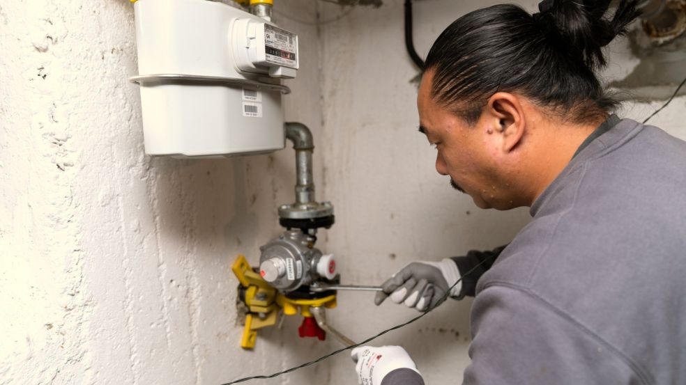 Loc Tran arbeitet in einem Kellerraum an einer Gasinstallation. Er trägt ein graues Langarmshirt und Handschuhe und führt mit einem langen Metallwerkzeug eine Einstellung an der Armatur unterhalb des Gaszählers durch.