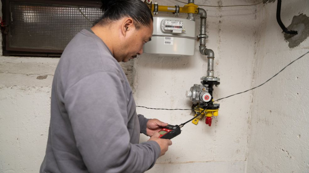 Loc Tran führt in einem Kellerraum eine technische Messung an einer Gasinstallation durch. Er trägt ein graues Langarmshirt und hält ein elektronisches Messgerät in den Händen, dessen Kabel zu einem Bauteil der Gasleitung führt. Vor ihm befinden sich der montierte Gaszähler. An der Wand ist zudem ein kleines Kellerfenster zu sehen, durch das gedämpftes Licht einfällt.