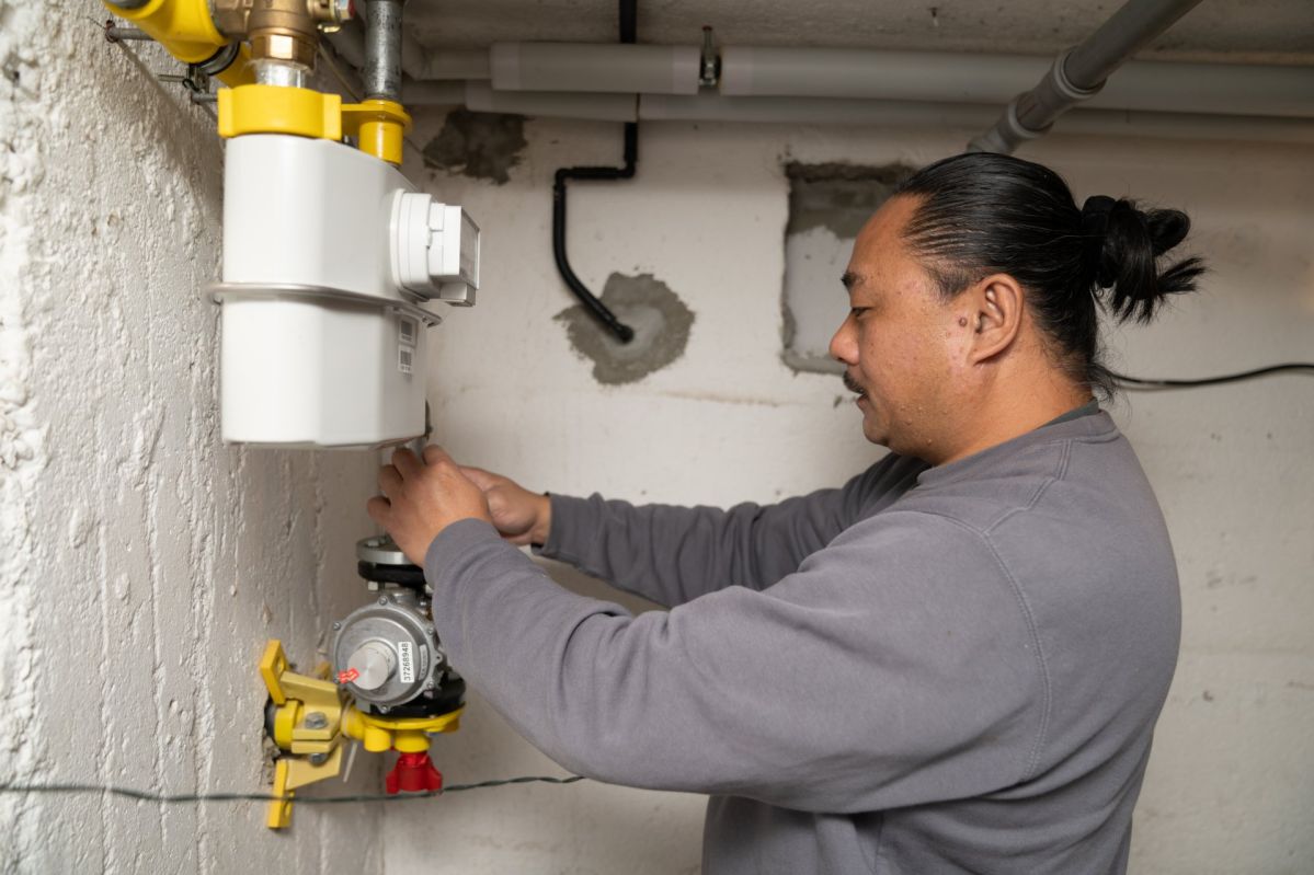 Loc Tran arbeitet in einem Kellerraum an einem Gaszähler. Er trägt ein graues Langarmshirt und steht seitlich vor einer weiß verputzten Wand. Vor ihm befindet sich die Installation mit Gaszähler, gelben Absperrhähnen und angeschlossenen Leitungen. Mit beiden Händen bedient er eine Komponente des Zählers, während oberhalb und entlang der Wand weitere Rohre verlaufen, die Teil der technischen Anlage sind.