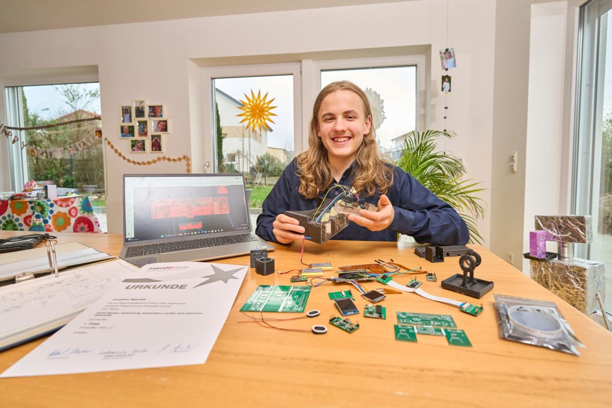 Jonathan und seine kleine Schwester Elina Baschek Eine Person sitzt an einem Tisch mit Elektronikkomponenten und hält ein Gerät. Ein Laptop zeigt steht daneben. Im Hintergrund sieht man ein Wohnzimmer mit Fenstern. Auf dem Tisch liegt ein Blatt mit "URKUNDE".