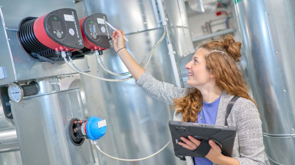 Energieeffizienz im Einsatz Valerie Schmitz bei der technischen Inspektion: Mit Tablet in der Hand prüft sie zwei rot-schwarze Geräte an einer großen metallischen Anlage.