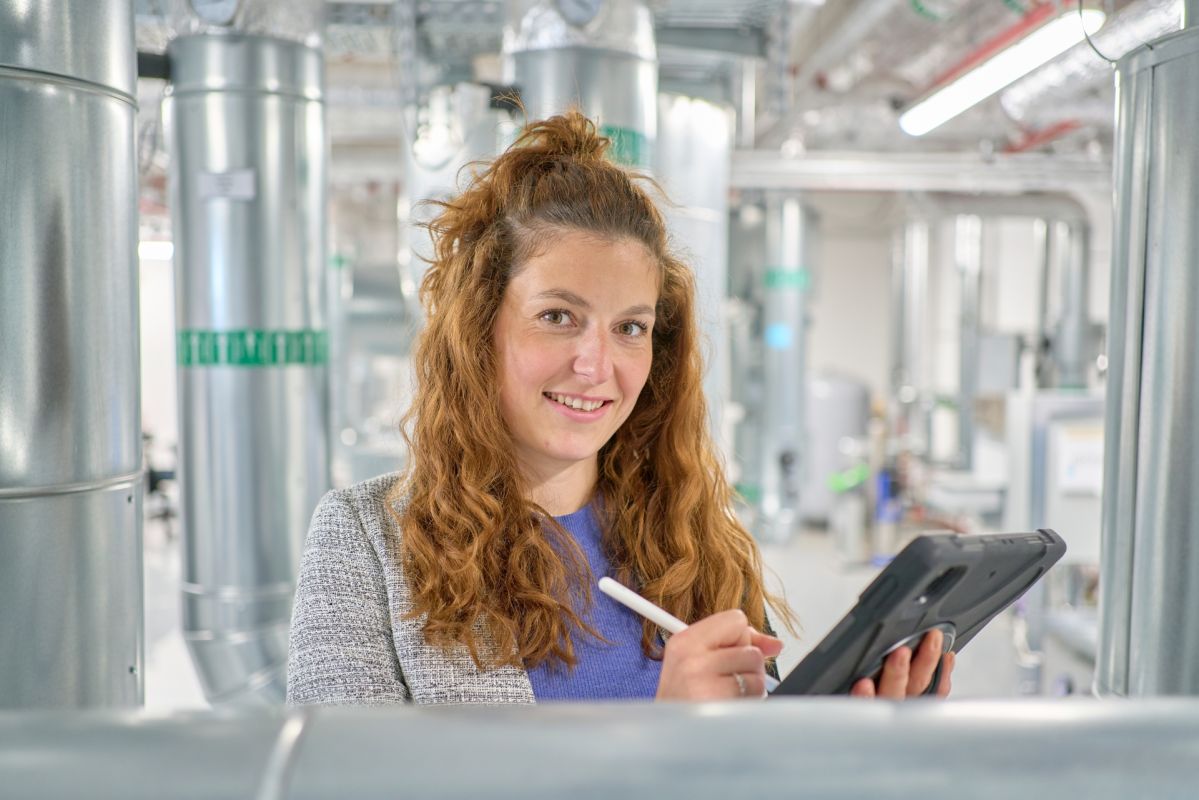 Energieeffizienz im Einsatz Valerie Schmitz bei der Arbeit: Mit dem Tablet dokumentiert sie vor Ort energetische Verbrauchsdaten. Im Hintergrund sind große Rohrleitungen und industrielle Anlagen zu sehen.