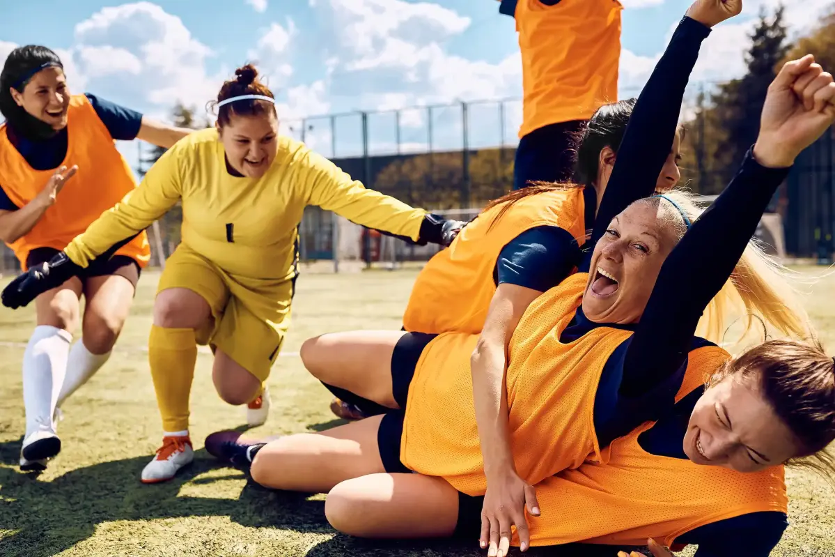 Ein Frauenfußballteam feiert ausgelassen auf einem Spielfeld im Freien. Die Spielerinnen tragen orangefarbene Leibchen und jubeln liegend auf dem Rasen. Im Hintergrund sonniger Himmel und Bäume.