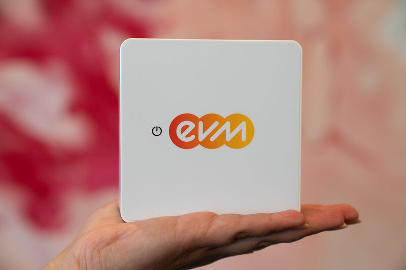 evm+ Energiepilot - Der Energiepilot für dein Zuhause Eine Hand hält ein quadratisches, weißes Gerät mit dem farbigen „evm“-Logo