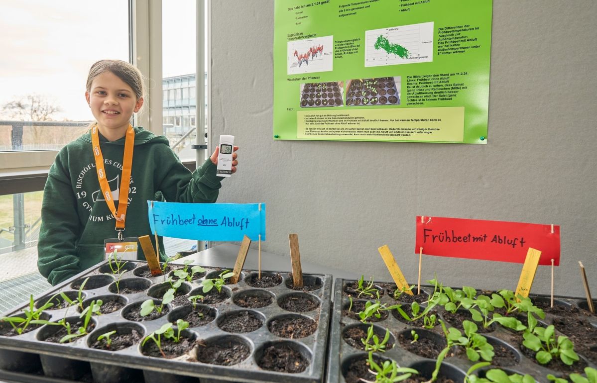 Elisa Baschek untersucht für Schüler experimentieren, wozu Luft, Licht und Wärme bei der Gemüsezucht gut sind Ein Kind hält ein Gerät und steht neben zwei Pflanzschalen mit Keimlingen, die Schilder "Frühbeet ohne Abwurf" und "Frühbeet mit Abwurf" tragen. Im Hintergrund ein Plakat.