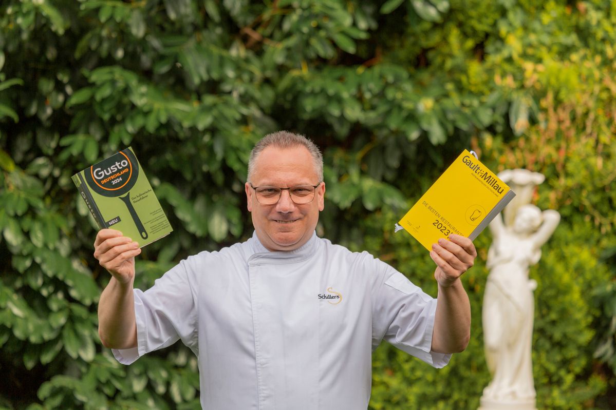 Ein Mann in einem weißen Kochoutfit hält zwei Bücher mit den Titeln "Gusto" und "Gault&Millau 2023" vor einem Hintergrund aus dichtem, grünem Laub und einer weißen Statue.