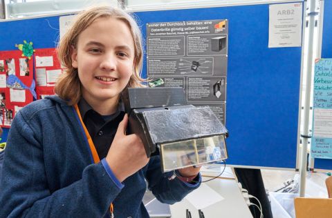 Datenbrille von Jonathan Baschek Eine Person hält ein selbstgebautes elektronisches Gerät vor einer Präsentationstafel in einer Ausstellungshalle. Auf der Tafel steht: "Innen den Durchblick behalten: eine Datenbrille günstig selber bauen".