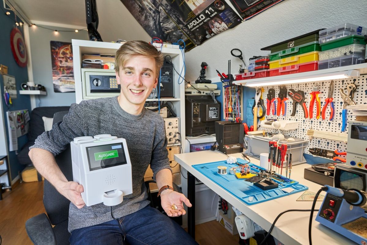 Jugend forscht 2022-Pillendreher Ein junger Mann hält einen selbstgebauten Automaten mit einem Display mit der Aufschrift „Ausgabe“ in einem Heimwerkstattzimmer voller Werkzeuge und Elektronikbauteile lächelnd in die Kamera.