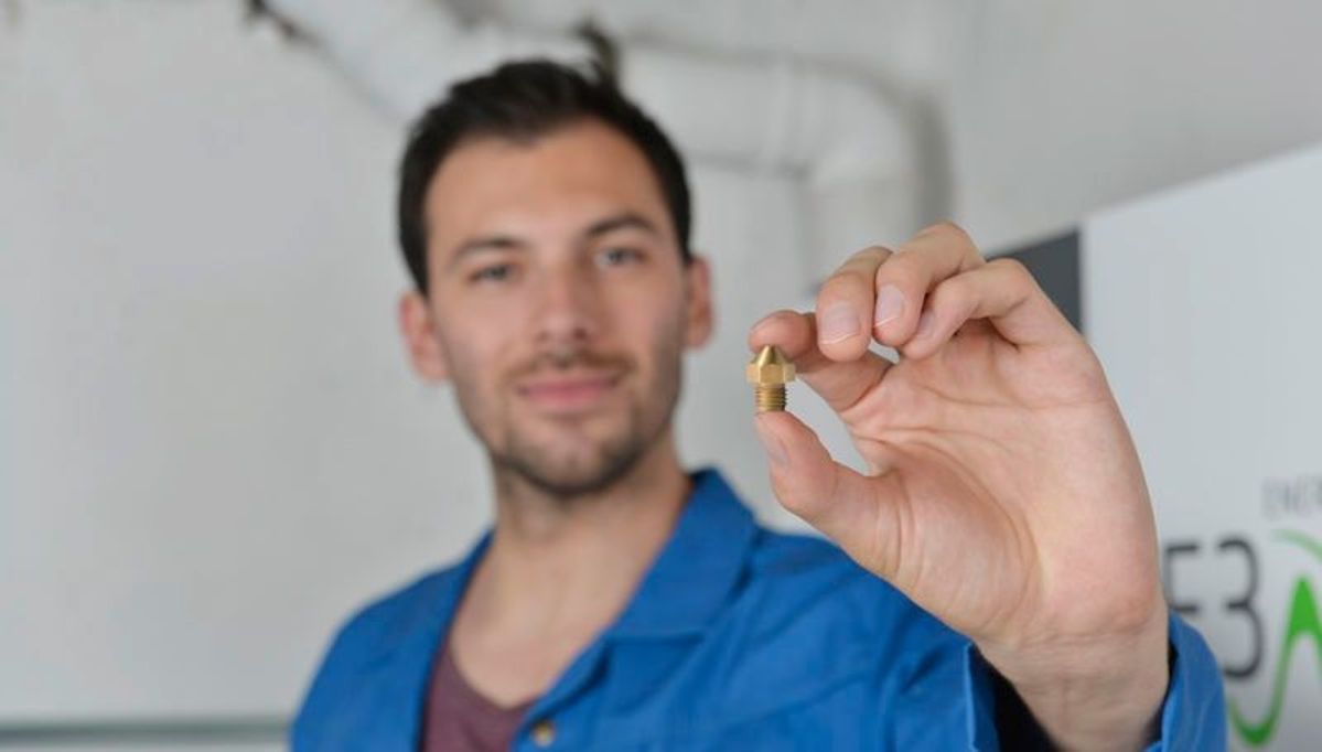 Ein Mann in einem blauen Hemd hält eine kleine Schraube zwischen Daumen und Finger, während er in einem Büro mit Rohrleitungen im Hintergrund steht.