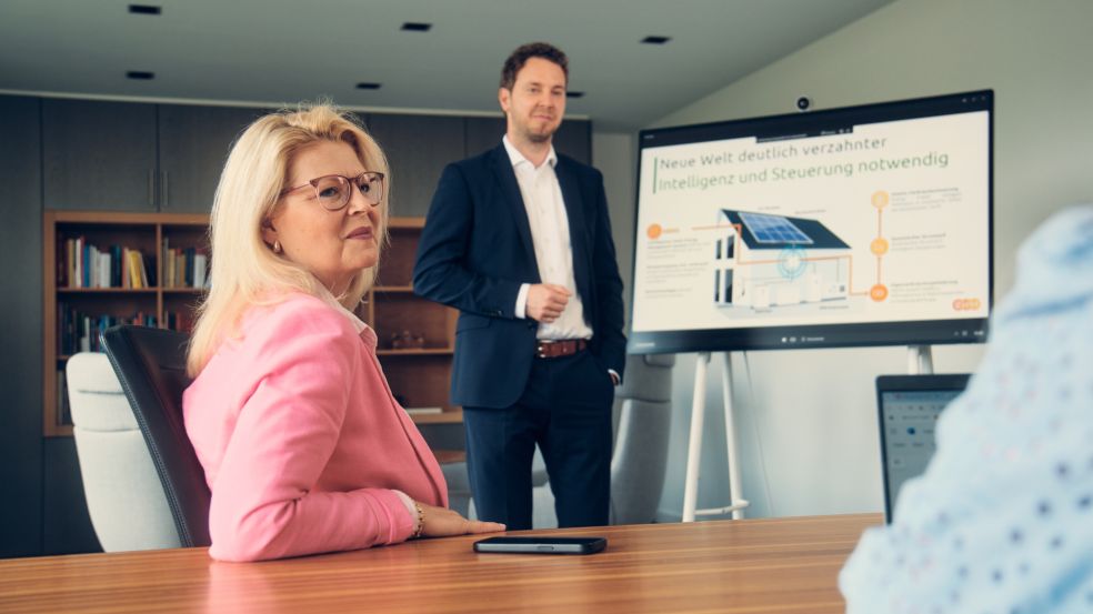 Nicole De Leuw sitzt an einem Tisch in einem Besprechungsraum. Im Hintergrund sieht man Christoph Hesse eine Präsentation halten.