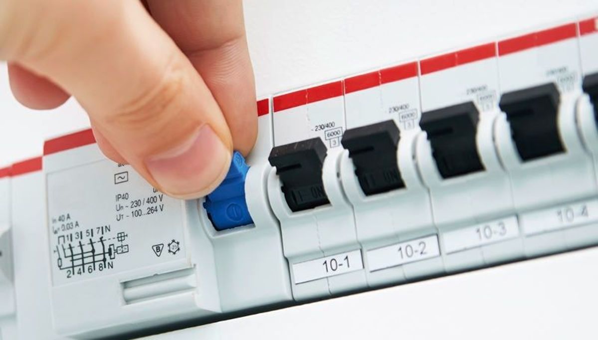 Eine Hand schaltet einen blauen Schalter in einem Sicherungskasten umgeben von mehreren schwarzen Schaltern um