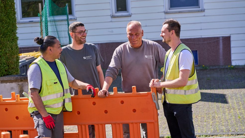 Vier Mitarbeitende der Energienetze Mittelrhein (enm) stehen in Arbeitskleidung mit Warnwesten und Handschuhen vor einer Baustelle. Im Hintergrund sind ein weißes Gebäude mit Fenstern und eine grün abgedeckte Struktur zu sehen.