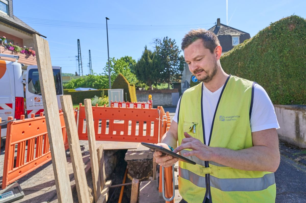 Ein Bauarbeiter der Energienetze Mittelrhein (enm) steht mit gelber Warnweste und Tablet vor einem offenen Graben, umgeben von Absperrbarken, Baumaterialien und Rohren. Im Hintergrund sind Gebäude, Bäume und Stromleitungen unter blauem Himmel zu sehen.