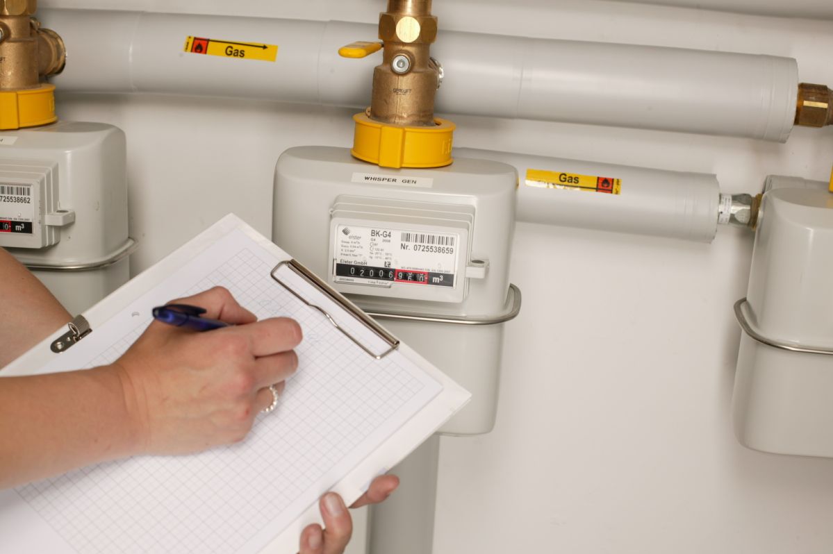 Eine Person notiert Zählerstände auf einem Klemmbrett vor zwei Gaszählern in einem Technikraum. Auf den Zählern steht "GAS METER" und "Gas."