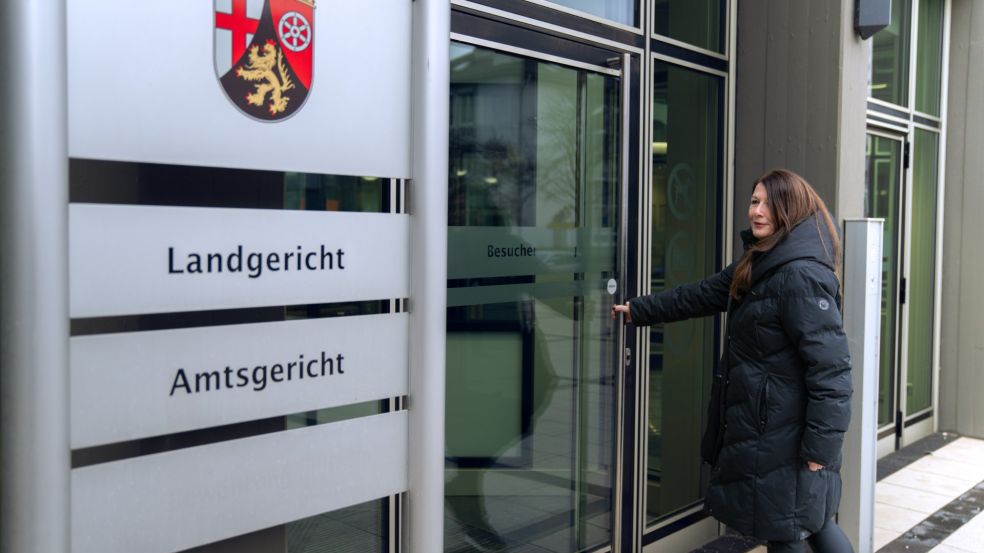 Silke Kras öffnet eine Glastür an einem modernen Gerichtsgebäude. Neben dem Eingang steht ein Schild mit dem Wappen von Rheinland-Pfalz sowie den gut lesbaren Bezeichnungen „Landgericht“ und „Amtsgericht“.