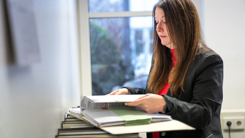 Silke Kras steht an einem hohen Ablage- oder Aktenschrank in einem Büro und arbeitet mit mehreren gestapelten Aktenordnern und Unterlagen. Sie hält ein geöffnetes Dokument in den Händen und blättert darin. Im Hintergrund ist ein Fenster zu sehen, durch das Tageslicht in den Raum fällt.