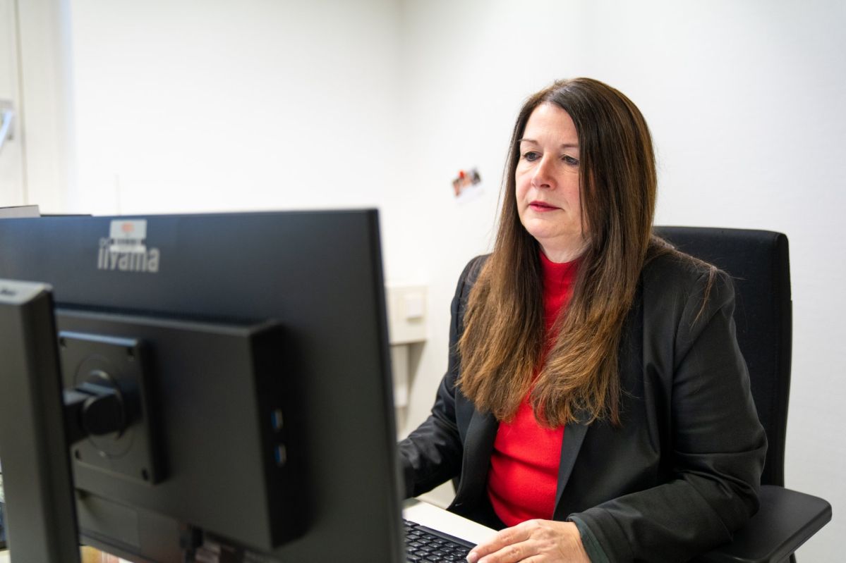 Silke Kras sitzt an einem Schreibtisch in einem Büro und arbeitet an einem Computer mit großem Monitor. Sie befindet sich auf einem Bürostuhl und hat die Hände auf Tastatur und Maus positioniert. Die Wand im Hintergrund ist hell, technische Anschlüsse sind sichtbar.