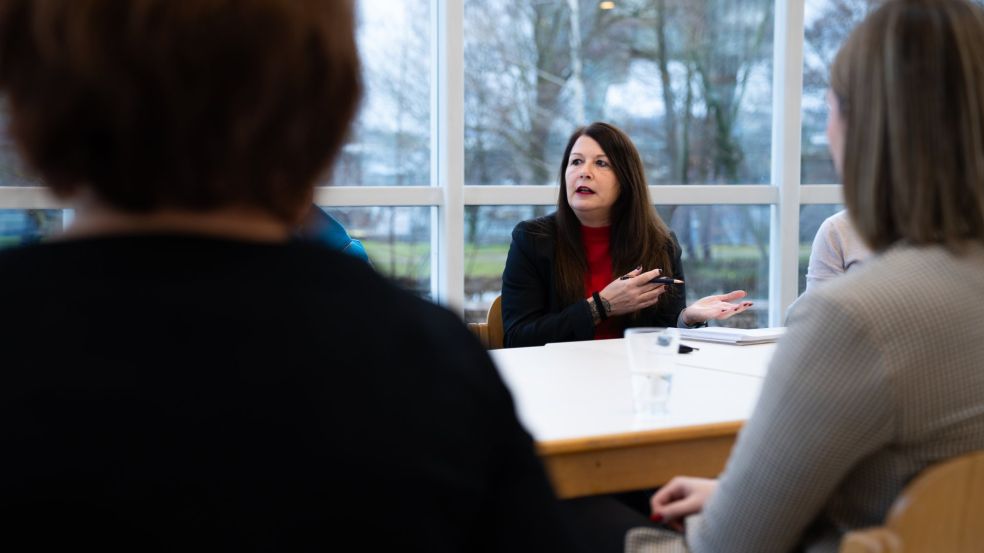 Silke Kras sitzt an einem Besprechungstisch und spricht zu mehreren Kolleginnen und Kollegen, die um den Tisch herum Platz genommen haben. Vor ihr liegen Unterlagen und ein Glas Wasser. Der Raum ist hell und verfügt über große Fenster mit Blick ins Freie.